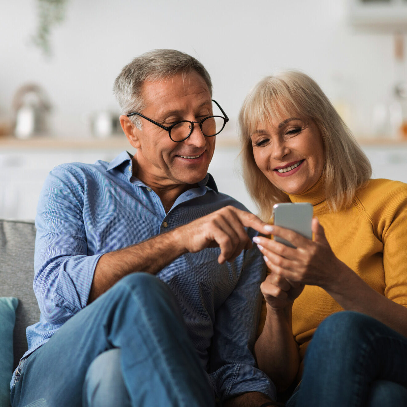 Older Spouses Using Mobile Phone Together, Pointing Finger At Cellphone Texting And Browsing Internet Sitting On Couch At Home. Mature Spouses Networking In Social Media Via Smartphone Application