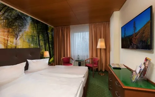 Hotelzimmer mit Doppelbett, Wandbild von Bäumen, TV, zwei Stühlen und Tisch.