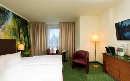 Hotelzimmer mit Doppelbett, Waldbild an der Wand, Sessel, TV und Fenster mit Vorhängen.
