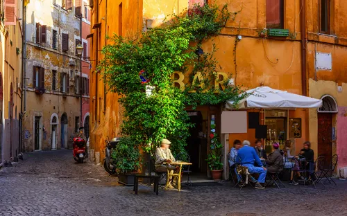 Gasse mit historischen Gebäuden, blauen Fensterläden und Außenterrasse bei Sonnenschein.