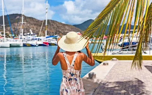 Frau mit Hut steht am Hafen, Boote und Palmen im Hintergrund.