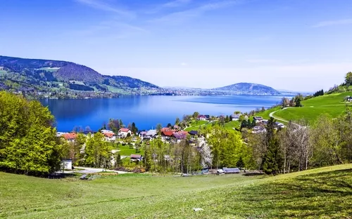 Landschaft mit See, Bergen und Dorf bei klarem Himmel.