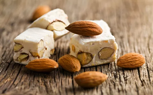 Weißer Nougat mit Mandeln auf rustikalem Holzuntergrund, umgeben von ganzen Mandeln.