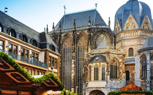 Bild des weihnachtlichen Katschhofs in Aachen, Nordrhein-Westfalen. Im Vordergrund befinden sich festlich geschmückte Marktstände, während im Hintergrund die historische Kathedrale mit ihrer mittelalterlichen Architektur beleuchtet zu sehen ist. Die Szene vermittelt eine traditionelle Weihnachtsstimmung im Advent.