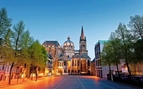 Blick auf den Aachener Dom und den Platz davor in der Abenddämmerung, umgeben von Bäumen und historischen Gebäuden.