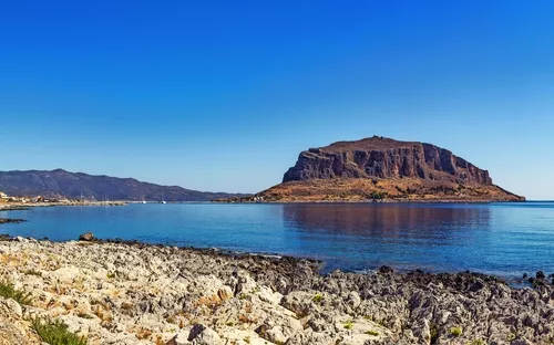 Felsige Küste mit großer Insel im Hintergrund und blauem Himmel.