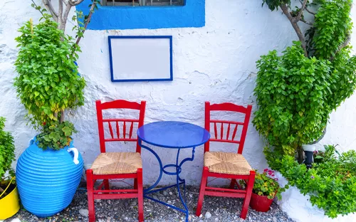 Zwei rote Stühle und ein blauer Tisch vor weißer Wand mit Pflanzen.