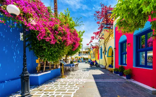 Bunte Gasse mit blühenden Bougainvilleen und Cafétischen im Freien.