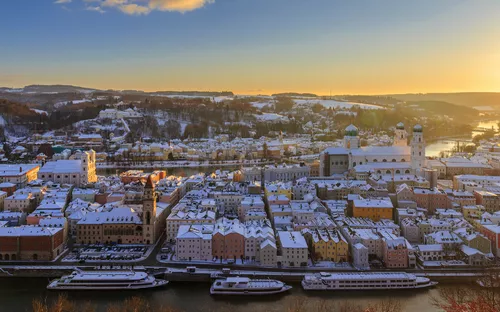 Verschneite Stadt an Flussufer bei Sonnenaufgang mit historischen Gebäuden.