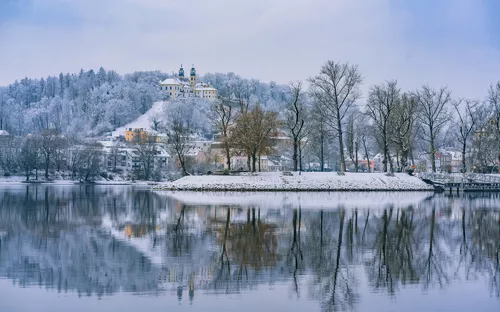 Verschneiter Hügel mit Kirche und Bäumen, gespiegelt in einem ruhigen See.
