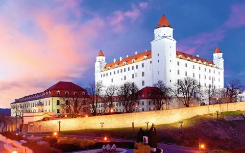 Schloss auf Hügel bei Sonnenuntergang, mit beleuchteten Mauern und rosa-blauem Himmel.