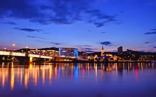 Stadtansicht bei Nacht mit beleuchteter Brücke und Gebäuden am Fluss.