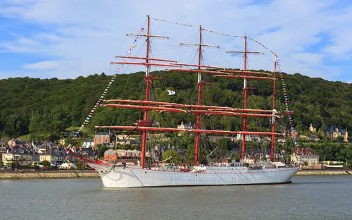 Dreimastiges Segelschiff auf Fluss vor grüner Hügellandschaft und Häusern.