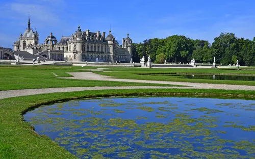 Schloss mit Garten und Teich im Vordergrund unter blauem Himmel.