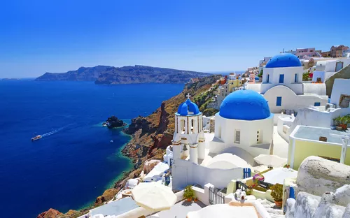 Weiße Gebäude mit blauen Kuppeln auf Santorini, Meer im Hintergrund.