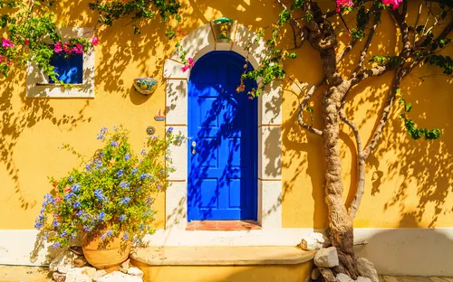 Blaue Tür an einem gelben Haus mit Blumentöpfen und Kletterpflanze im Sonnenlicht.