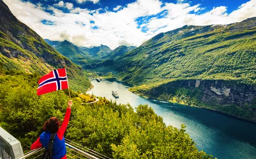 Person schwenkt norwegische Flagge vor Fjordlandschaft.