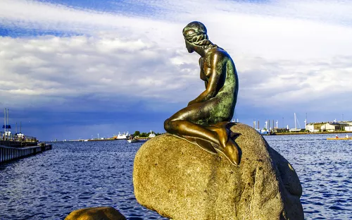 Skulptur einer Meerjungfrau auf einem Felsen am Wasser bei blauem Himmel.