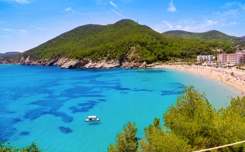 Strand mit türkisblauem Wasser, umgeben von grünen Hügeln und Hotels, mit einem Boot im Wasser.