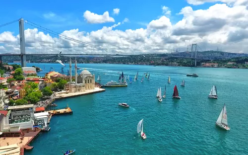 Blick auf die Bosporus-Brücke in Istanbul mit Segelbooten im Vordergrund und einer Moschee am Ufer.