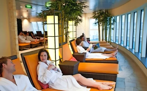 Personen entspannen auf Liegestühlen in einem Spa-Bereich mit Pflanzen und großen Fenstern.