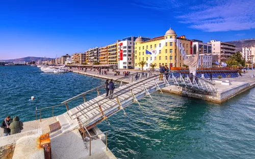 Hafenpromenade mit Fußgängerbrücke und Gebäuden am Wasser bei blauem Himmel.