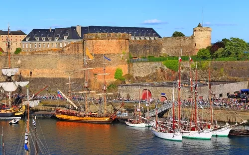 Alte Festung am Wasser, umgeben von vielen Segelbooten und Menschenmengen bei klarem Himmel.