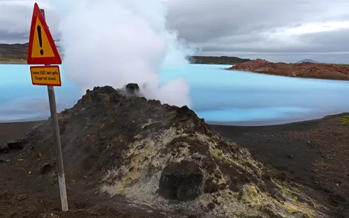 Vulkanische Landschaft mit dampfendem Boden und Warnschild nahe eines Gewässers.