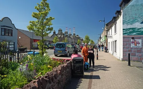 Belebte Straße mit Fußgängern, Autos und blühenden Blumen am Gehweg.