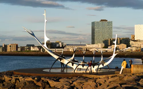 Metallskulptur am Ufer mit einer Stadt im Hintergrund bei Sonnenuntergang.