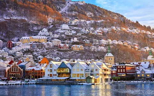 Winterlandschaft mit bunten Häusern am Wasser und schneebedeckten Hügeln im Hintergrund.