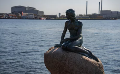 Bronzestatue einer Meerjungfrau auf einem Felsen im Wasser mit Industriekulisse.