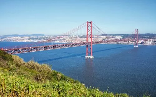 Rote Hängebrücke über einem Fluss mit Stadt im Hintergrund an einem klaren, sonnigen Tag.