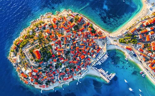 Luftbild einer Küstenstadt mit roten Dächern und Yachthafen am blauen Meer.