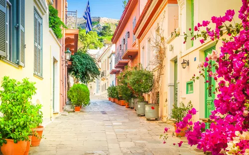 Bunte Gasse mit Blumen und griechischer Flagge im Hintergrund.