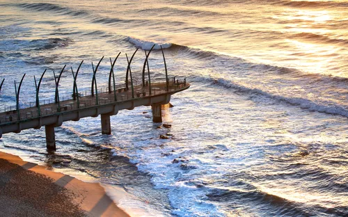 Pier, der ins Meer ragt, Ansicht bei Sonnenuntergang mit brechenden Wellen am Strand.