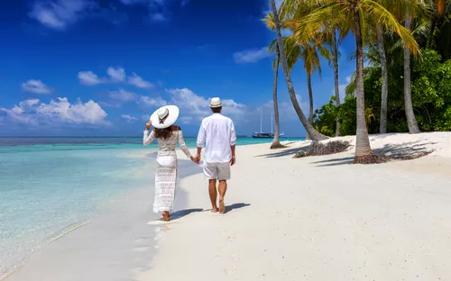 Paar spaziert am tropischen Sandstrand entlang, umgeben von Palmen und klarem Wasser.