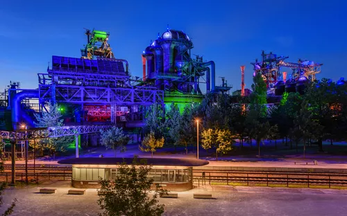 Beleuchtetes Industriegebäude bei Nacht mit Bäumen im Vordergrund.