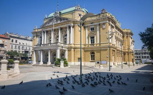 Historisches Gebäude mit Tauben auf dem Vorplatz bei blauem Himmel.