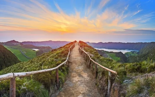 Wanderweg auf einem Hügel mit Sonnenuntergang und weitem Blick über Landschaft und See.