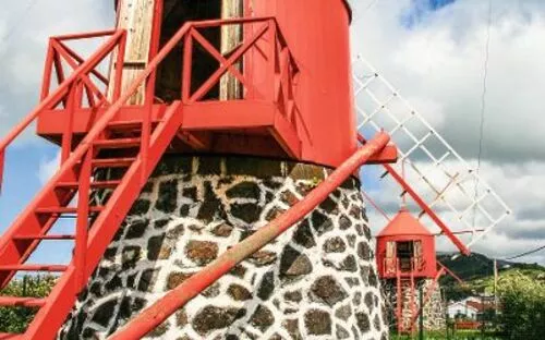Rote Windmühle mit Steinsockel auf grünem Rasen unter wolkigem Himmel.