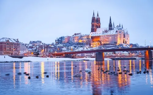 Schneebedeckte Burg und Stadtansicht mit Fluss im Vordergrund bei Dämmerung.