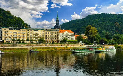 Ein Fluss mit einem Boot, im Hintergrund ein Gebäude und bewaldete Hügel.
