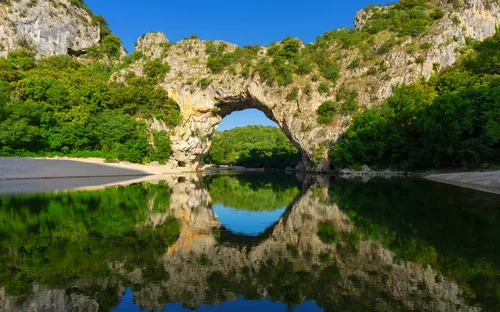 Natursteinbogen mit Spiegelung in ruhigem Gewässer, umgeben von Bäumen und Felsen.