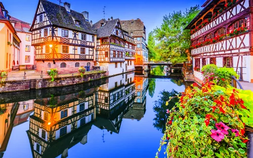 Fachwerkhäuser am Wasser mit Blumen im Vordergrund und spiegelnder Wasserfläche.