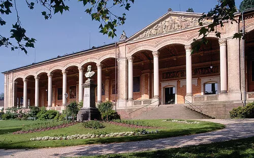 Historisches Gebäude mit Säulengang und Garten im Vordergrund.