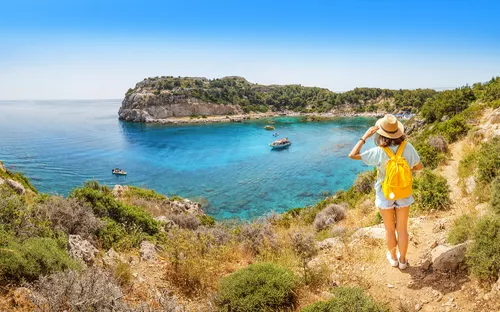 Frau mit gelbem Rucksack betrachtet malerische Küstenlandschaft mit türkisblauem Meer.