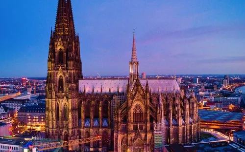 Kölner Dom bei Abenddämmerung mit beleuchtetem Weihnachtsmarkt davor.