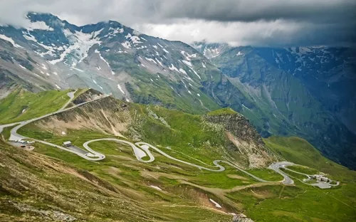 Serpentinenstraße durch grüne Berge mit schneebedeckten Gipfeln im Hintergrund.