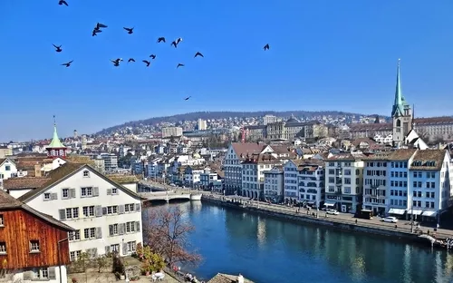 Stadtlandschaft von Zürich mit Häusern am Flussufer, einem Kirchturm und Vögeln am Himmel.